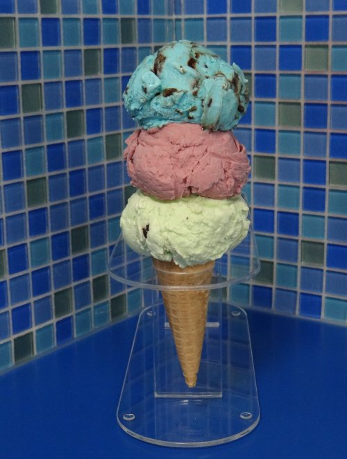 A tall ice cream cone with three scoops: blue mint chocolate chip, pink strawberry, and green pistachio, on a blue tiled background.