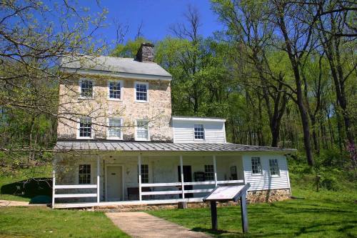 A historic two-story stone and wood house surrounded by lush green trees under a clear blue sky.