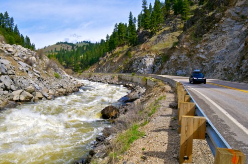 A winding road alongside a rushing river, surrounded by rocky cliffs and lush green trees under a clear blue sky.