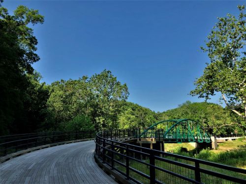 A winding wooden path leads to a green bridge over a serene river, surrounded by lush trees and a clear blue sky.