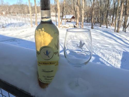 A bottle of Blue Heron Wines Traminette and a glass sit on a snowy railing, with a winter landscape in the background.