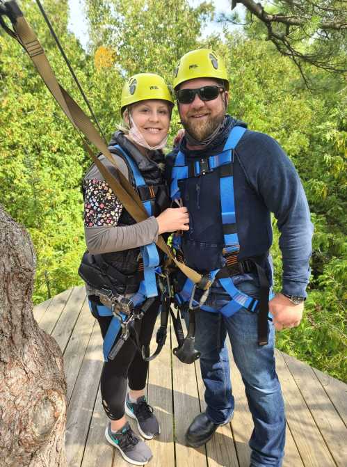 A couple in harnesses and helmets, smiling on a wooden platform surrounded by greenery, ready for ziplining.