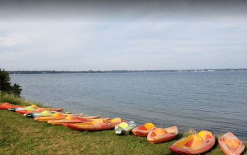 Colorful kayaks lined up on the grass by a calm lake under a cloudy sky.