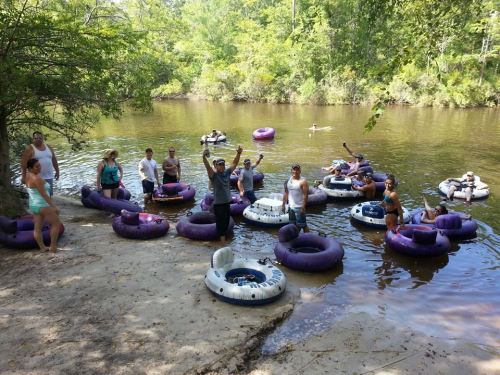 A group of people enjoying a sunny day floating on inner tubes in a river, surrounded by trees.