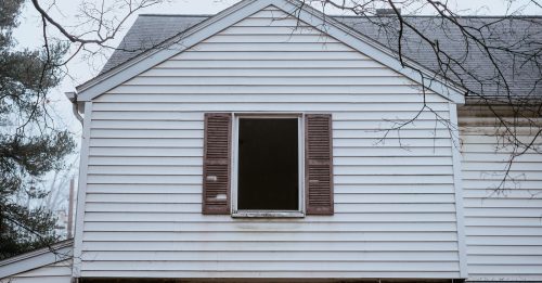 A close-up of a house's upper facade featuring a window with brown shutters and a bare tree branch in the foreground.