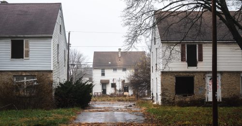 Abandoned houses line a overgrown street, with a foggy atmosphere and overcast sky in the background.