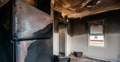 A charred kitchen with a blackened refrigerator and soot-covered walls, showing signs of fire damage and neglect.