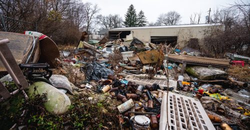 A cluttered area filled with debris, trash, and overgrown vegetation near a building on a cloudy day.