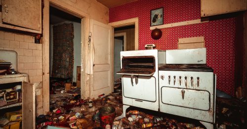 An abandoned kitchen filled with clutter, featuring an old stove and peeling wallpaper.