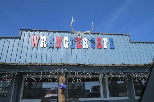 Sign for Wrangler Cafe with colorful letters and deer antlers on a blue building under a clear sky.