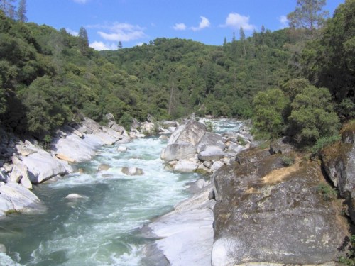 A flowing river surrounded by lush greenery and rocky banks under a clear blue sky.