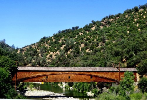 A rustic covered bridge spans a river, surrounded by lush green hills under a clear blue sky.