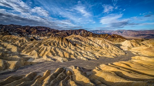 A panoramic view of rugged, layered desert terrain under a blue sky with scattered clouds.