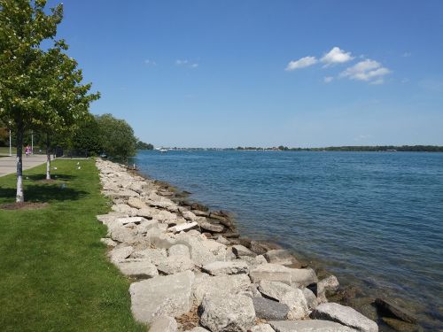 A serene lakeside view with rocky shore, green trees, and clear blue water under a partly cloudy sky.