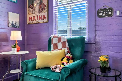 Cozy purple room with a teal armchair, decorative pillows, a lamp, and a small table with flowers and a penguin plush toy.