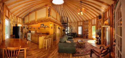 Cozy interior of a yurt with wooden beams, a kitchen, dining area, and comfortable seating. Bright and inviting atmosphere.