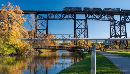 A train crosses a tall bridge over a river, surrounded by autumn trees and a walking path.