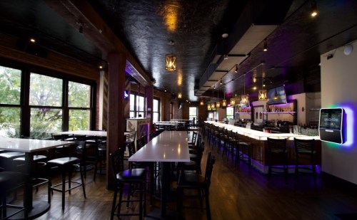 A modern bar interior with high tables, chairs, and a well-lit bar area featuring colorful lighting.