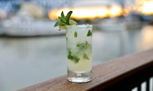 A refreshing cocktail with mint and lime, served in a tall glass, with a scenic waterfront background at sunset.