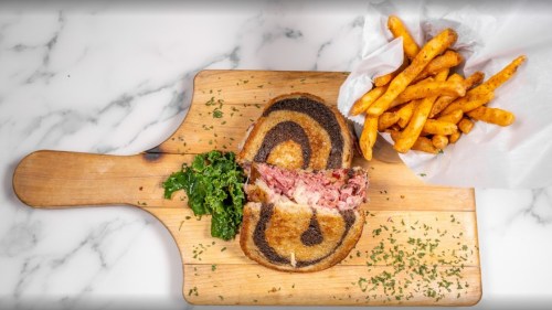 A sliced sandwich on a wooden board, featuring marbled bread, alongside sweet potato fries and a side of greens.