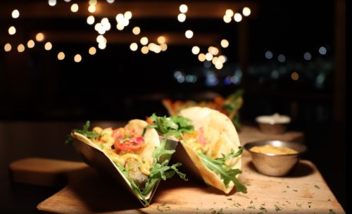 Two tacos filled with colorful ingredients on a wooden board, with dipping sauces and twinkling lights in the background.