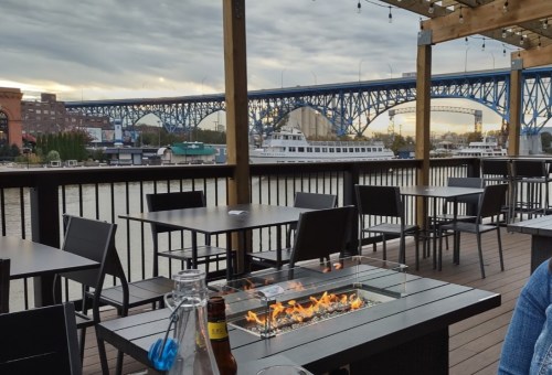Outdoor dining area with a fire pit, overlooking a river and a bridge under a cloudy sky.