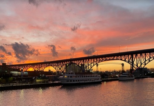 A vibrant sunset casts colors over a bridge and boats on a calm river, with clouds scattered in the sky.