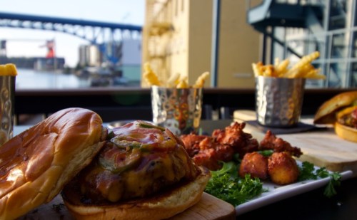 A close-up of a burger with cheese and jalapeños, fries in the background, and a scenic waterfront view.