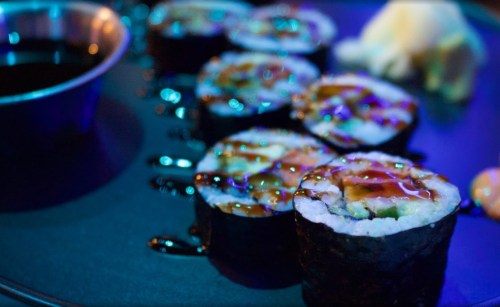 A close-up of sushi rolls drizzled with sauce, served on a dark plate with a small bowl of soy sauce.