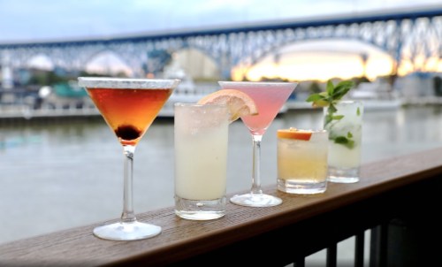 Five colorful cocktails on a wooden railing, with a river and bridge in the background during sunset.