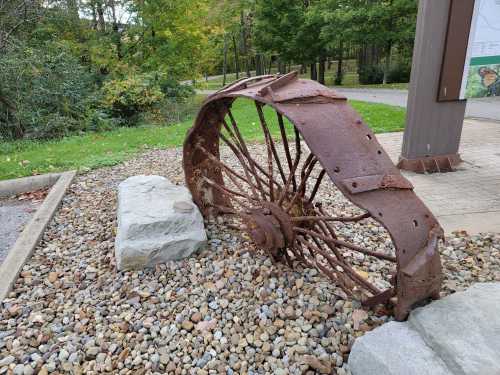 An old, rusted wheel partially embedded in gravel, surrounded by greenery and a pathway in the background.