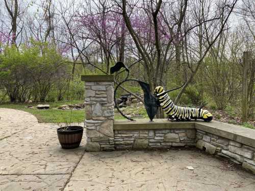 A whimsical garden scene featuring a large caterpillar sculpture beside a stone wall and a bird figure, surrounded by greenery.