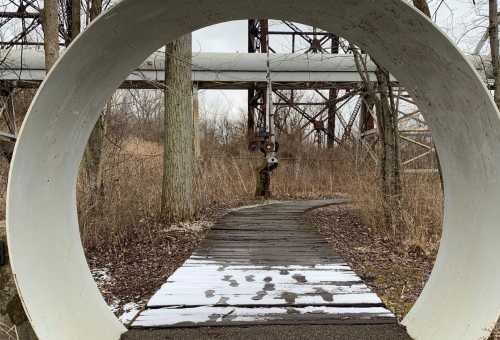 A circular concrete frame views a snowy path leading through overgrown grass and trees, with industrial structures in the background.