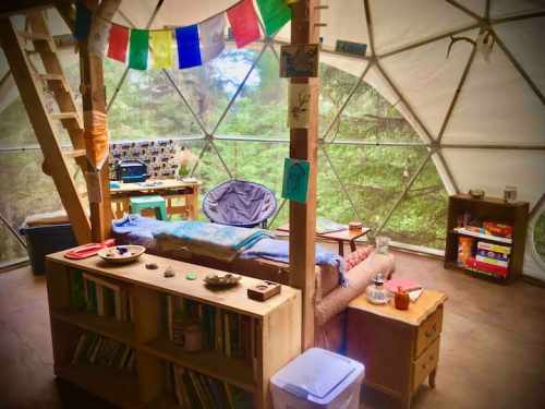 Cozy interior of a geodesic dome with a bed, bookshelf, and colorful decorations, surrounded by trees.