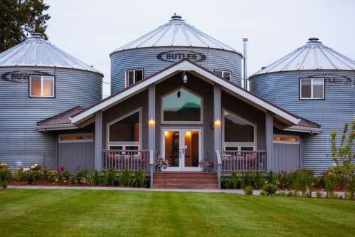 A modern home with a central entrance, flanked by two silos, surrounded by a green lawn and colorful flowers.