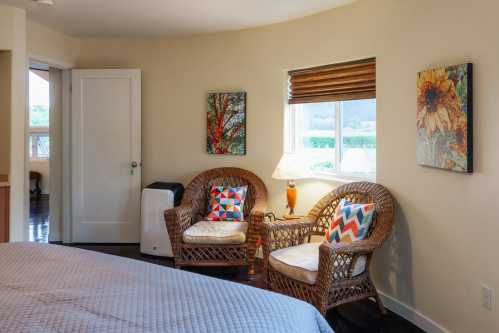 Cozy bedroom corner with two wicker chairs, colorful cushions, a lamp, and artwork on the walls.