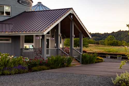 A charming farmhouse with a metal roof, surrounded by greenery and flowers, set against a scenic landscape.