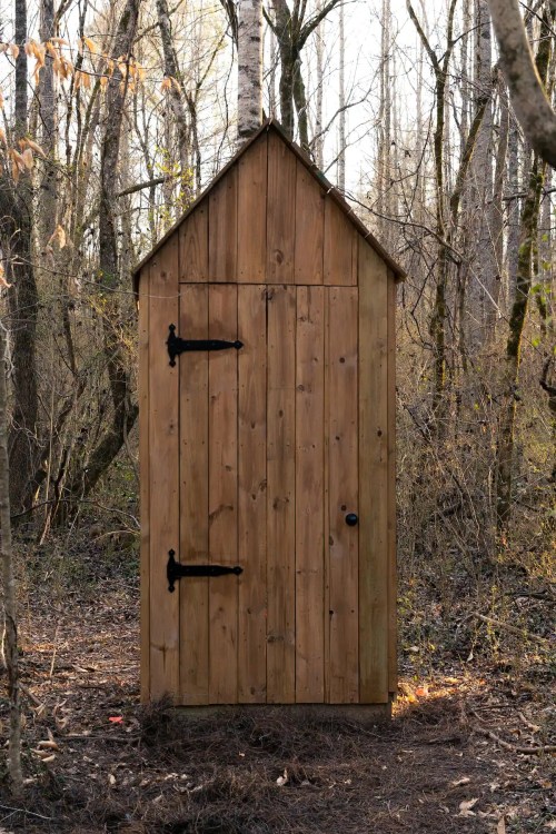 A rustic wooden outhouse stands alone in a wooded area, surrounded by trees and underbrush.