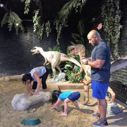 Two children and an adult interact with dinosaur models in a sandy play area surrounded by greenery.