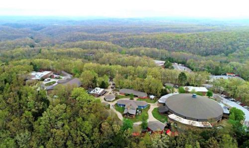 Aerial view of a wooded area with buildings nestled among trees, surrounded by rolling hills and greenery.