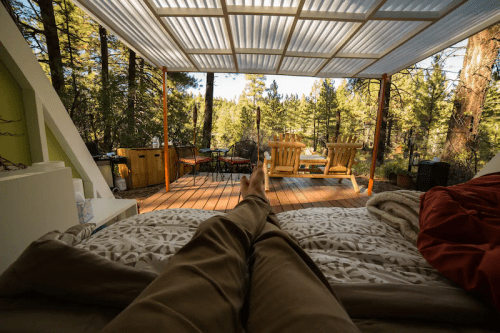 A person relaxes on a bed, looking out at a forest view from a cozy, covered deck with wooden furniture.