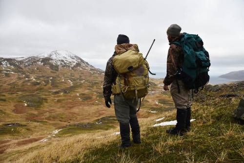 Two hikers with large backpacks stand on a grassy hill, overlooking a mountainous landscape under a cloudy sky.