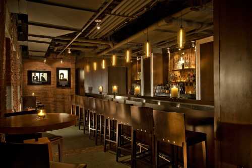 A dimly lit bar with wooden stools, a sleek counter, and shelves of liquor, creating a cozy atmosphere.