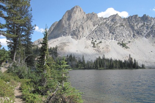 A serene lake surrounded by tall trees and a rugged mountain peak under a clear blue sky.