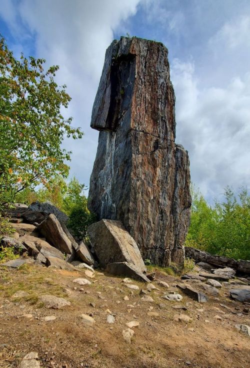 A tall, vertical rock formation surrounded by greenery and scattered stones under a partly cloudy sky.