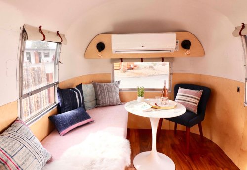 Cozy interior of a camper with a round table, chairs, and decorative pillows, featuring a small air conditioner.