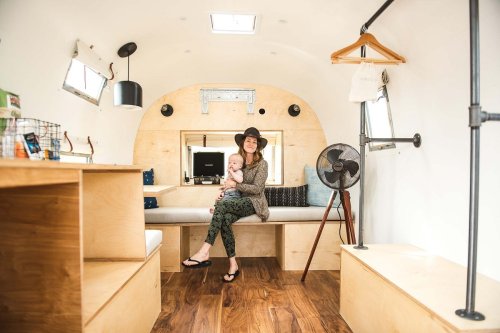 A woman in a hat holds a baby inside a cozy, modern camper with wooden accents and a minimalist design.