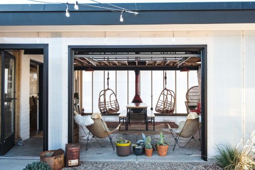 A cozy outdoor seating area with hanging chairs, a central table, and potted plants, framed by a modern structure.