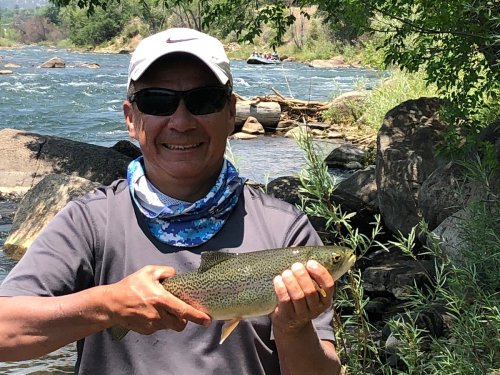 A man in sunglasses holds a rainbow trout by a river, surrounded by greenery and rocky terrain.