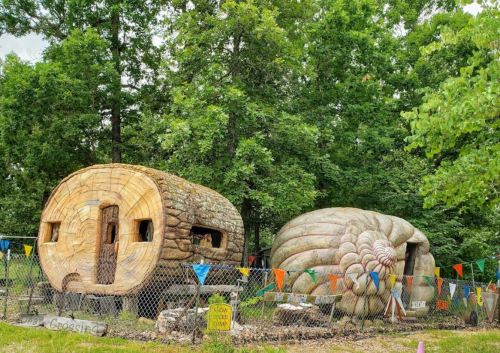 Two whimsical, oversized wooden structures resembling a log cabin and a giant snail, surrounded by trees and colorful flags.